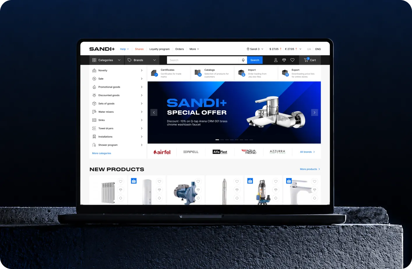 Laptop on a stone pedestal showing the redesigned Sandi+ B2B catalog home with a special-offer banner and new products